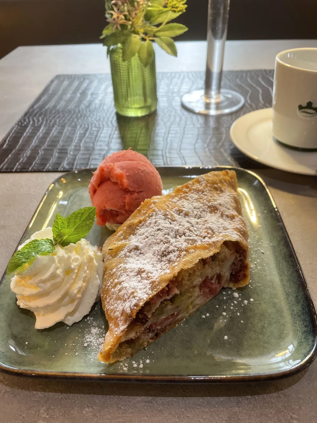 Strudel mit Puderzucker auf gr&uuml;nem Teller, dazu eine Kugel Sorbet, Schlagsahne und Minze, im Hintergrund eine Blumenvase