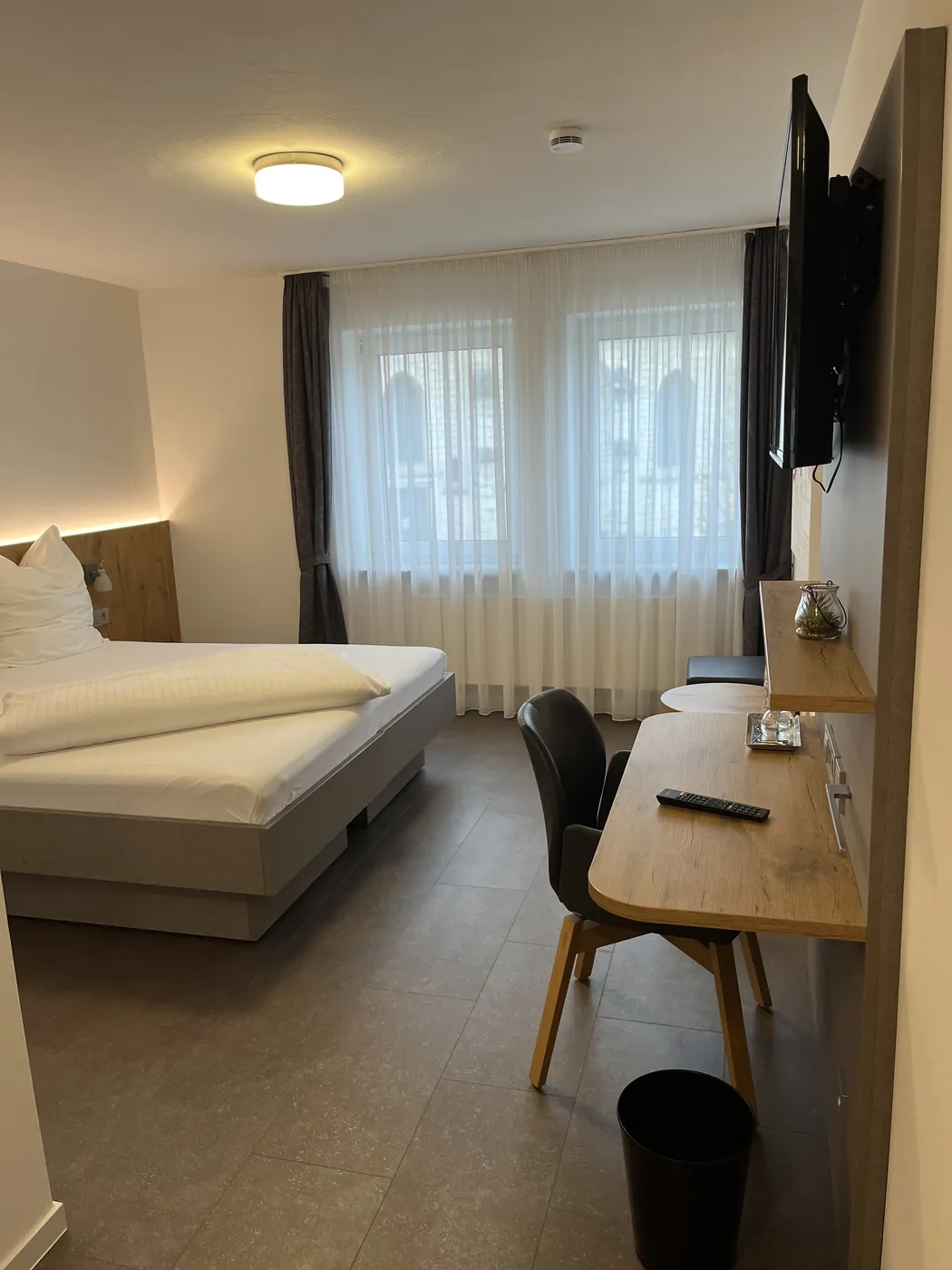 Zimmer mit bodengleichem Fliesenboden, Bett ohne Aufbau, Schreibtisch mit Stuhl und zwei Fenstern